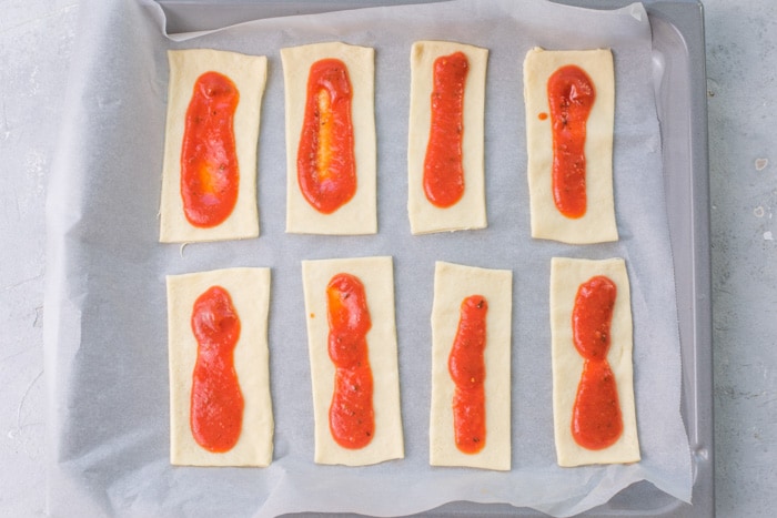 Rectangles of pizza dough topped with pizza sauce on a piece of parchment paper.