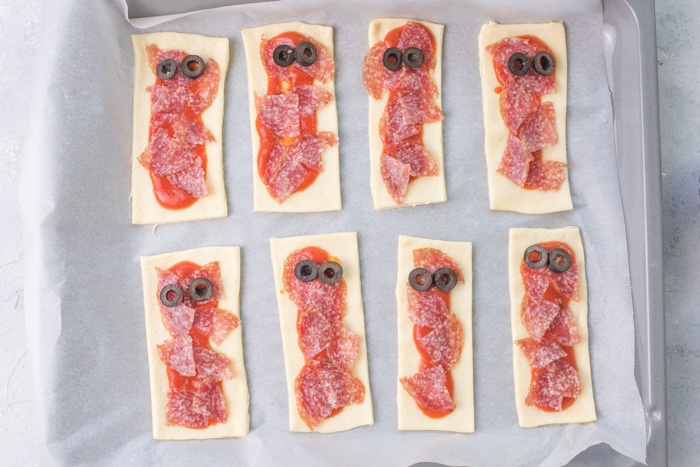 Rectangles of pizza dough topped with pizza sauce, salami, and olives.