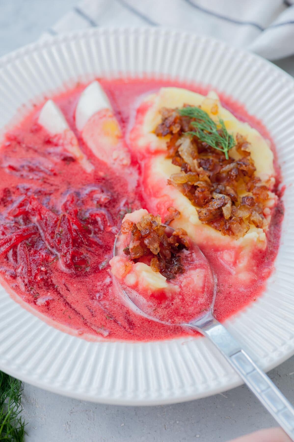 Barszcz soup in a white plate with mashed potatoed and onion bacon topping.