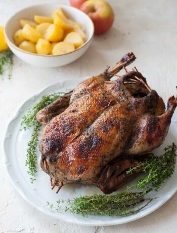 Roast duck on a white plate. Apples and potatoes in the background.