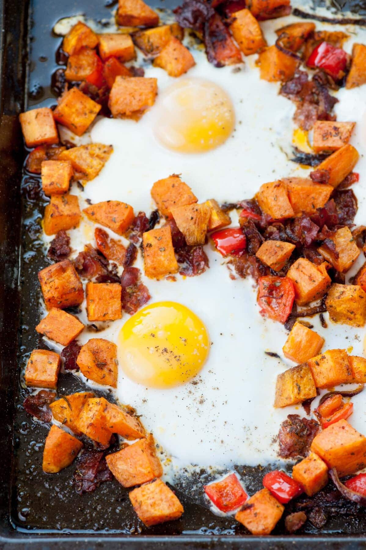 Sweet potato hash with bacon, bell, peppers and eggs on a black baking sheet.