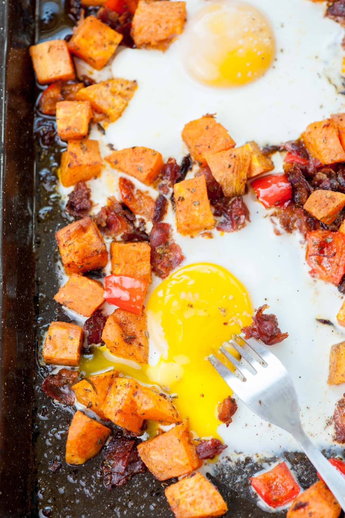 Sweet potato hash with bacon, bell, peppers and eggs on a black baking sheet. Fork is poking runny egg yolk.