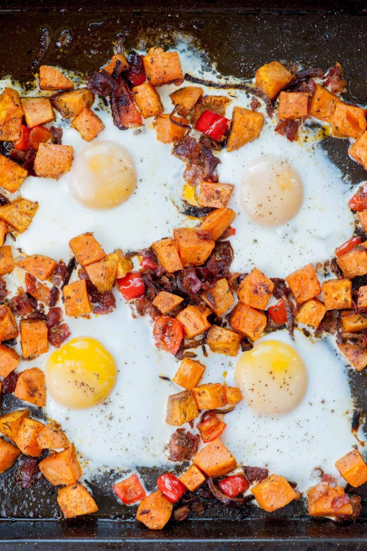 Sweet potato hash with bacon, bell, peppers and eggs on a black baking sheet.