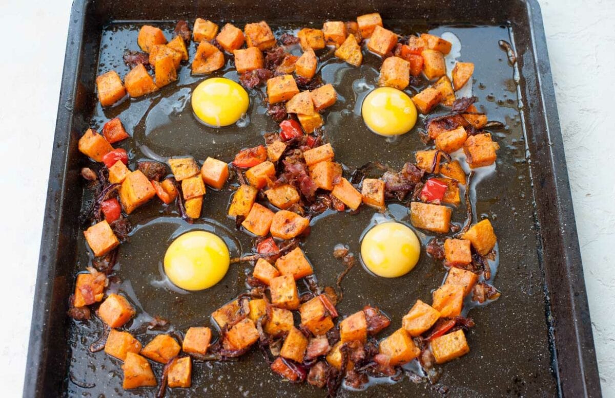 Sweet potato hash with bacon, bell, peppers and eggs ready to be baked on a black baking sheet.