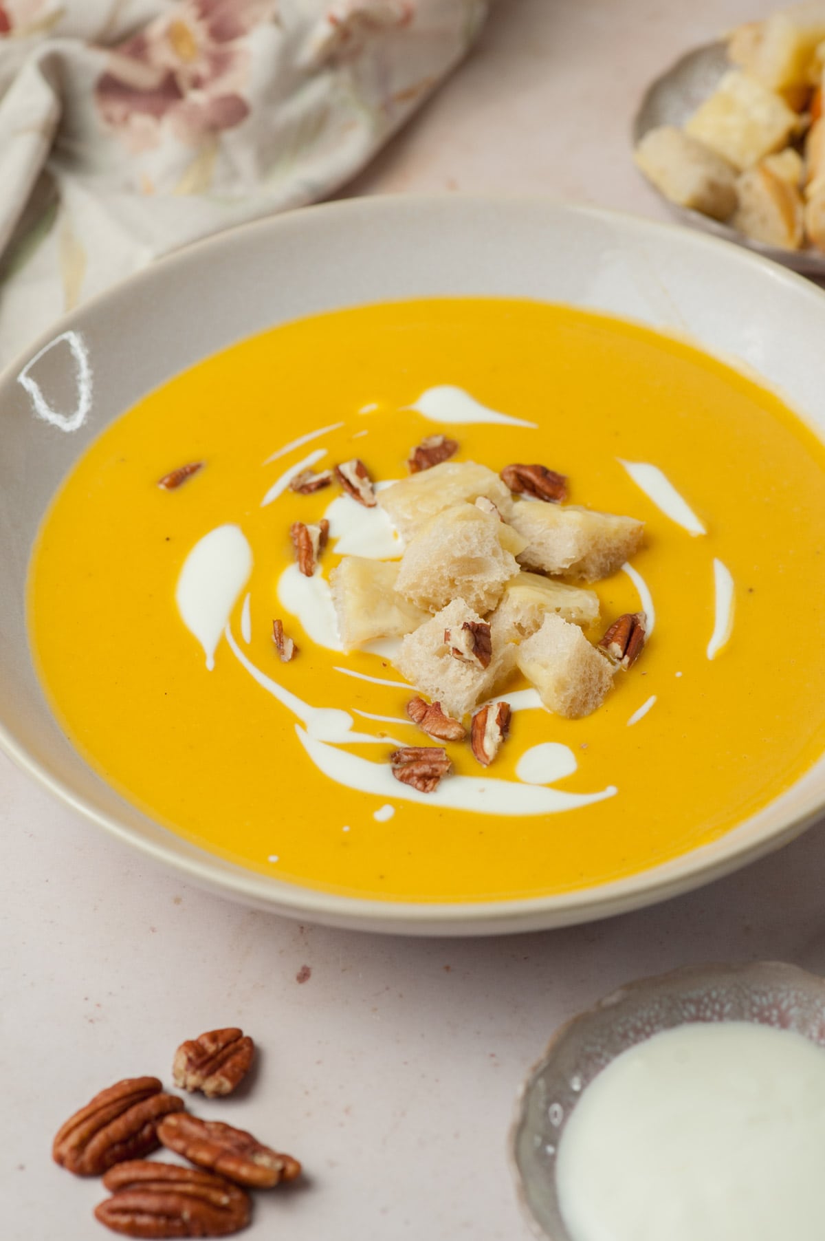 Butternut squash soup in a beige bowl topped with cheese croutons, pecans, and swirls of yogurt.