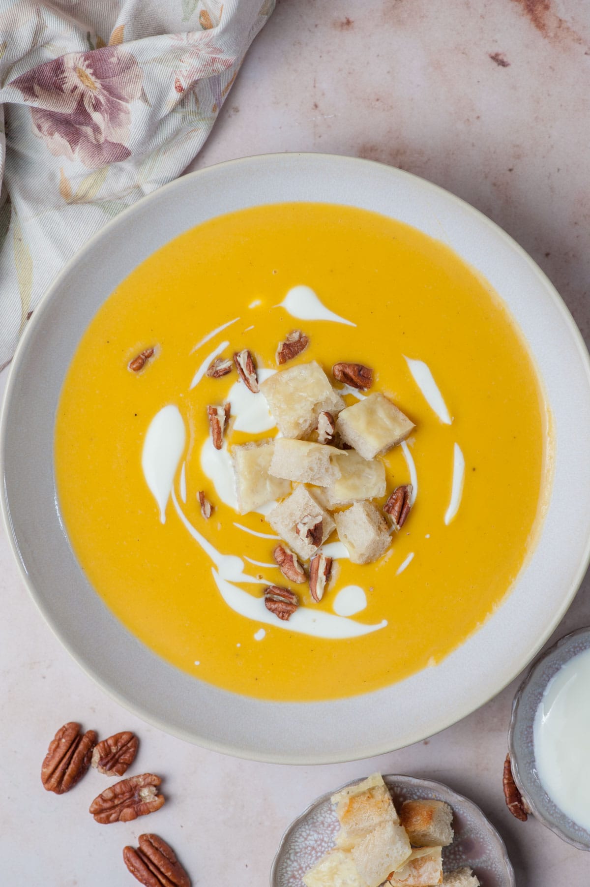 An overhead photo of butternut squash soup in a beige bowl topped with cheese croutons, pecans, and swirls of yogurt.