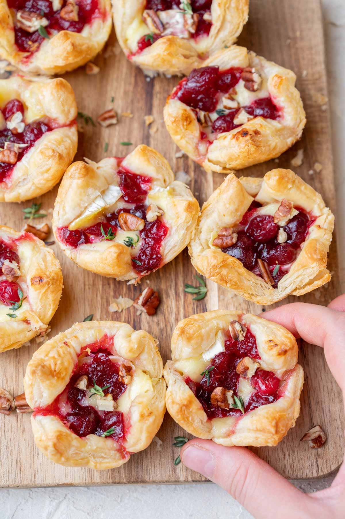 Puff pastry brie cranberry bites on a wooden board. One pastry is held in a hand.