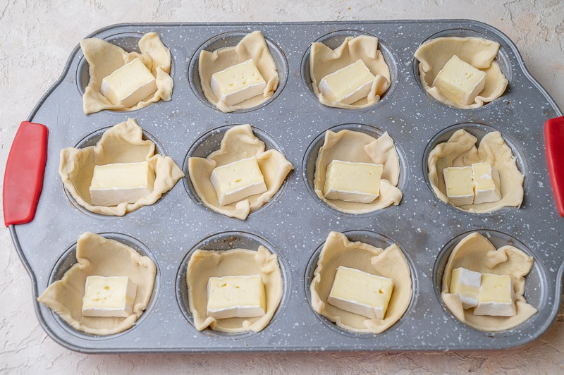 Puff pastry squares in a muffin tray with cubes of brie cheese.