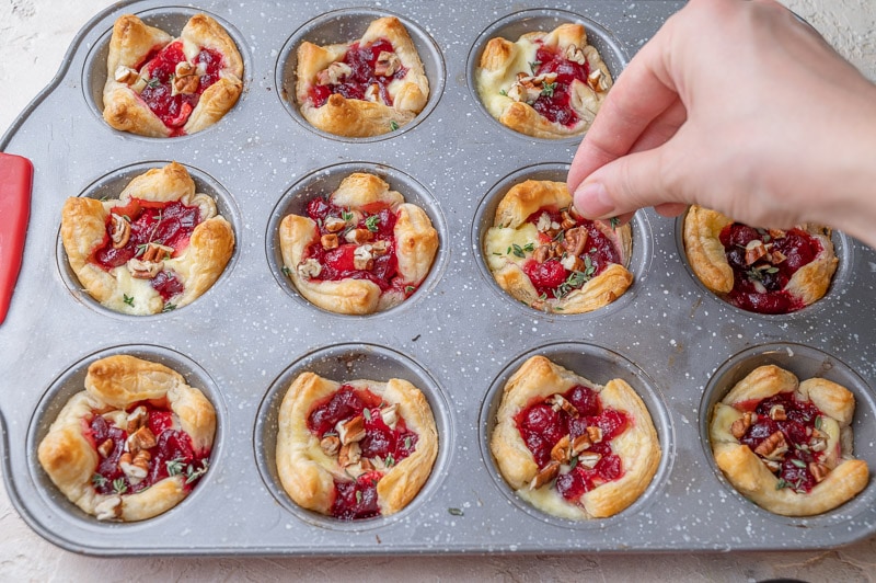 Baked puff pastry bites with brie and cranberry sauce are being sprinkled with pecans and thyme leaves.