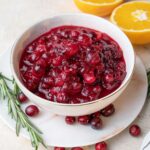 Cranberry orange sauce in a white bowl. Oranges and rosemary twigs in the background.