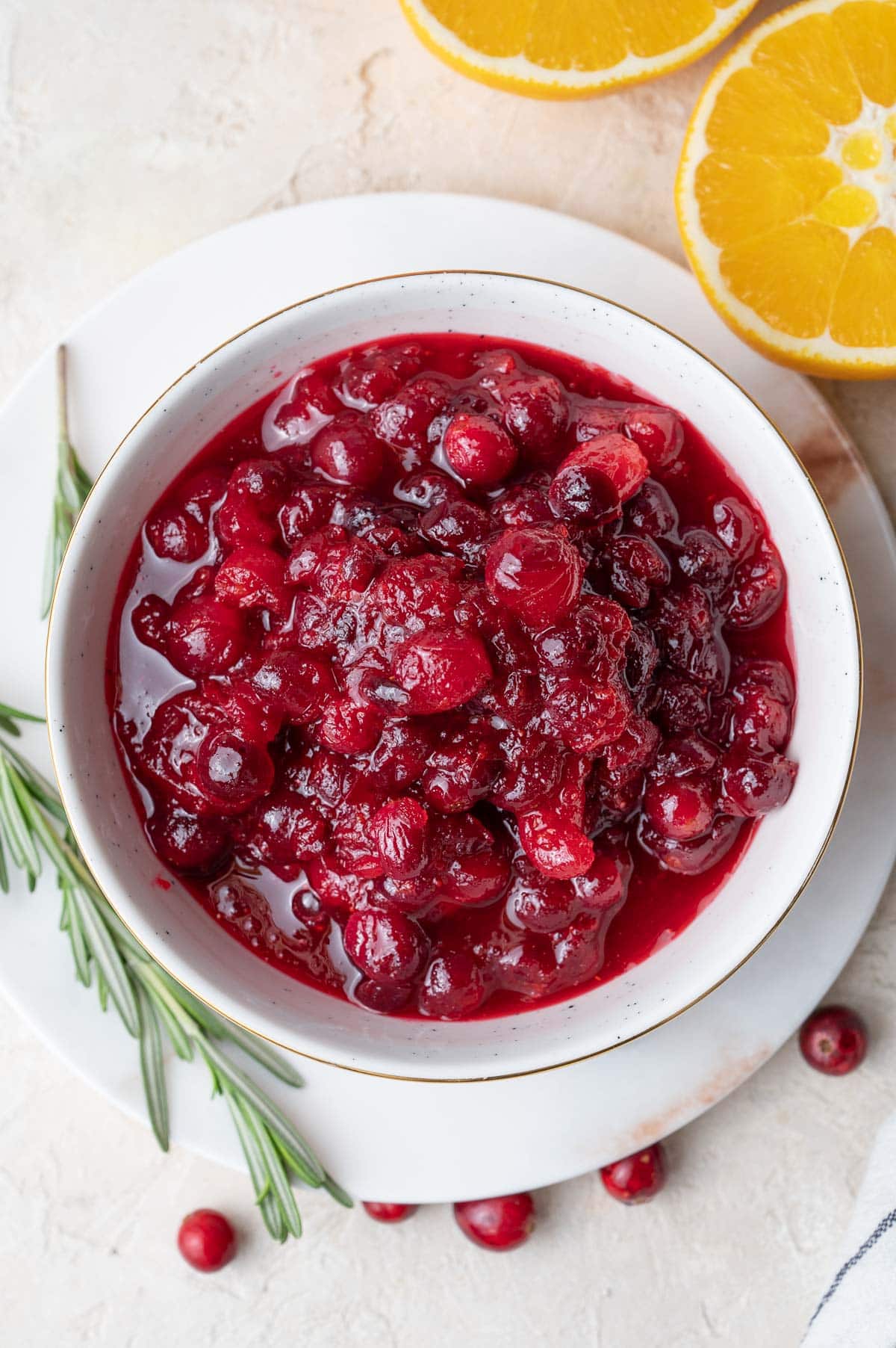 An overhead photo of cranberry orange sauce in a white bowl. Oranges and rosemary twigs in the background.