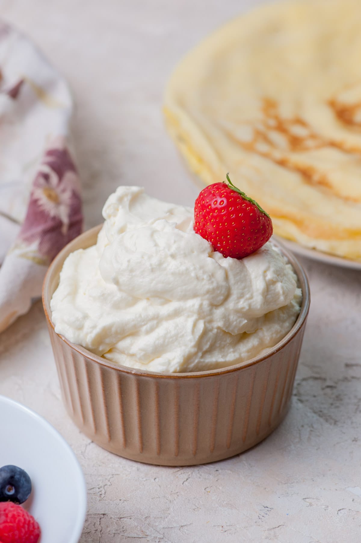 Cream cheese crepe filling in a small brown bowl topped with a strawberry. Crepes on a plate in the background.