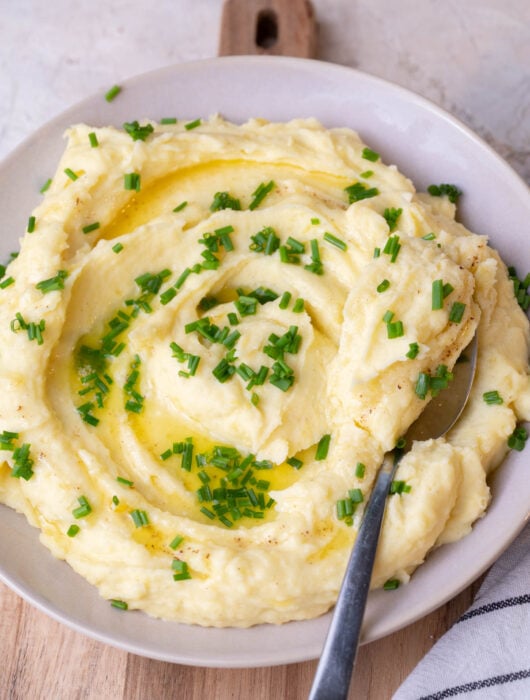 Cream cheese mashed potatoes in a beige bowl topped with chives, a spoon on the side.