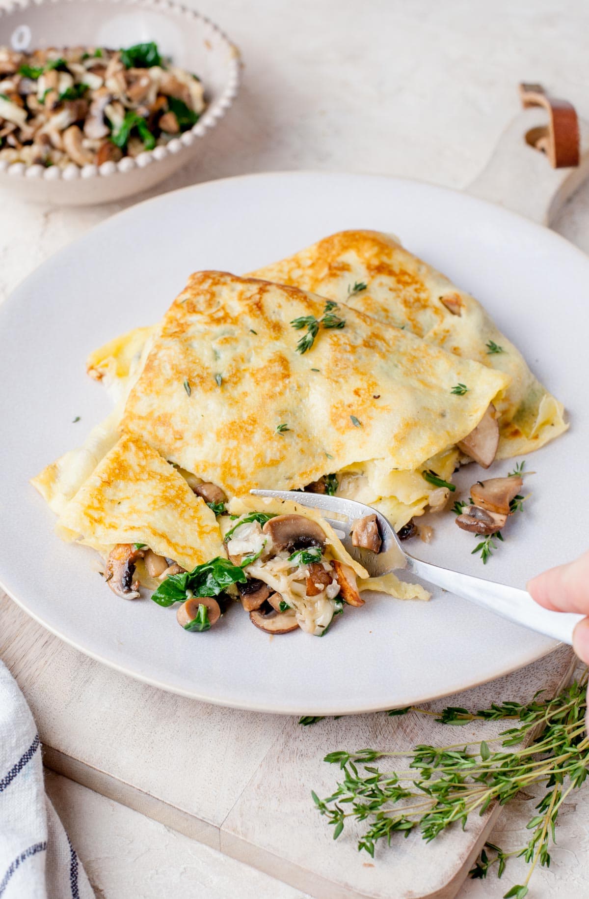 Two mushroom filled crepes in a beige plate are being cut with a fork.