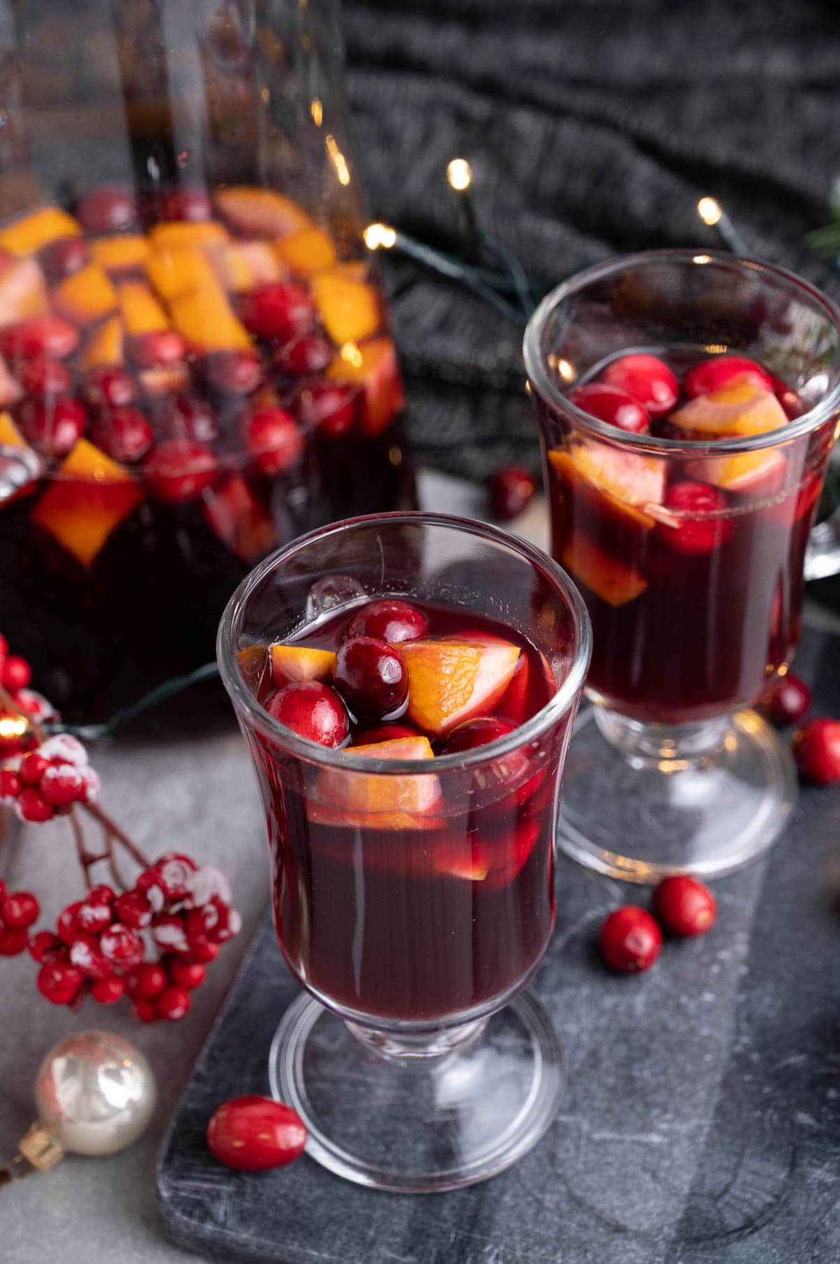 Two glasses with Christmas sangria on a black board. A pitcher and Christmas lights in the background.