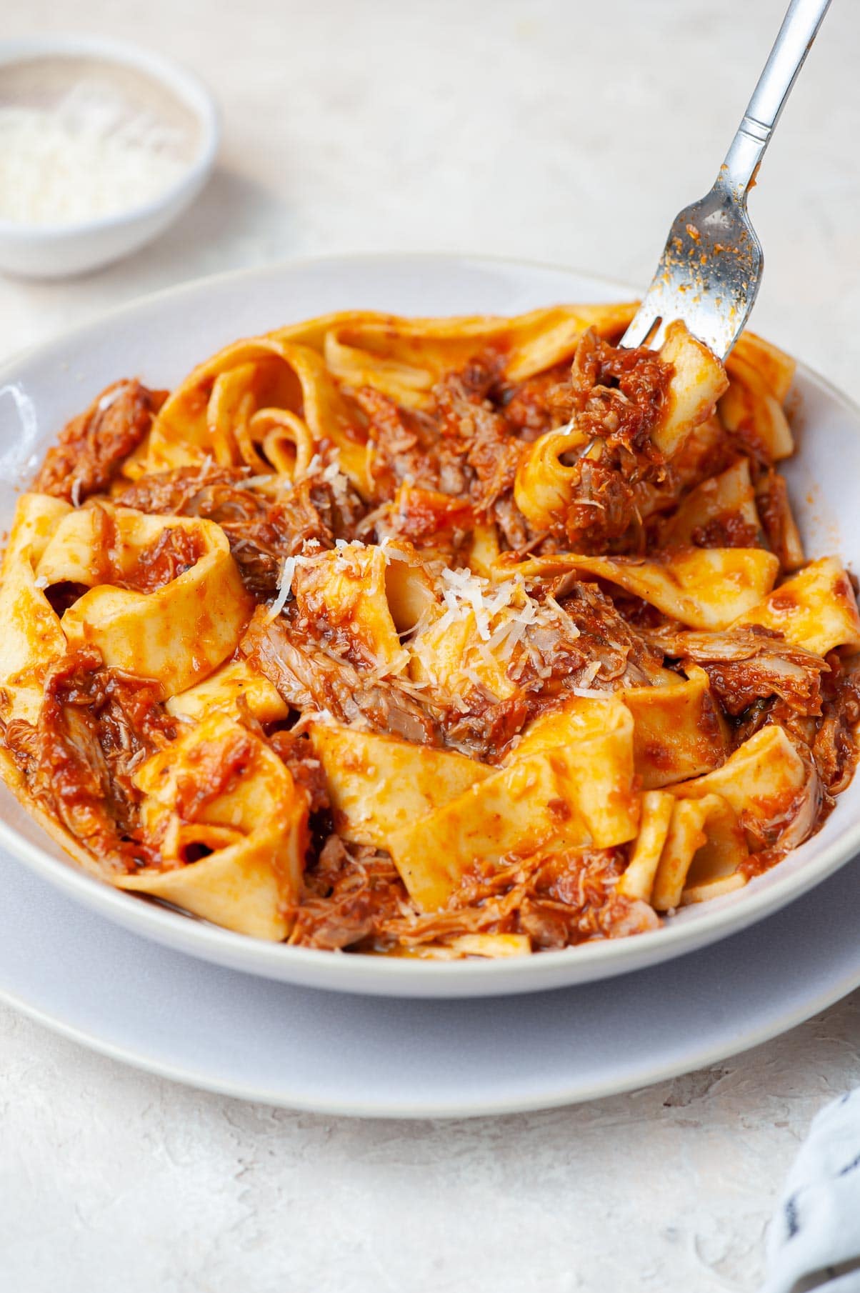 Duck ragu with pappardele pasta in a beige plate is being stuck on a fork.