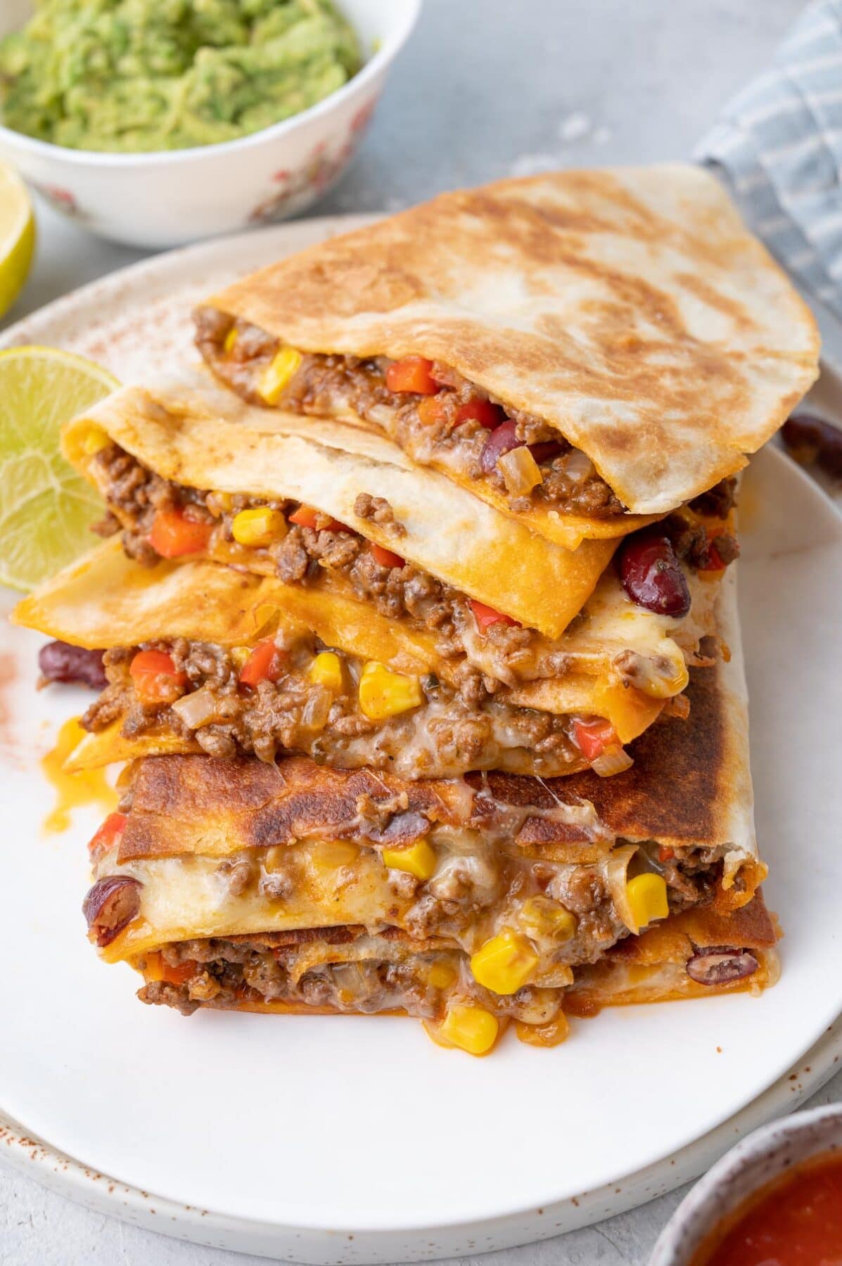 A stack of ground beef quesadillas in a white plate. Limes and guacamole in the background.