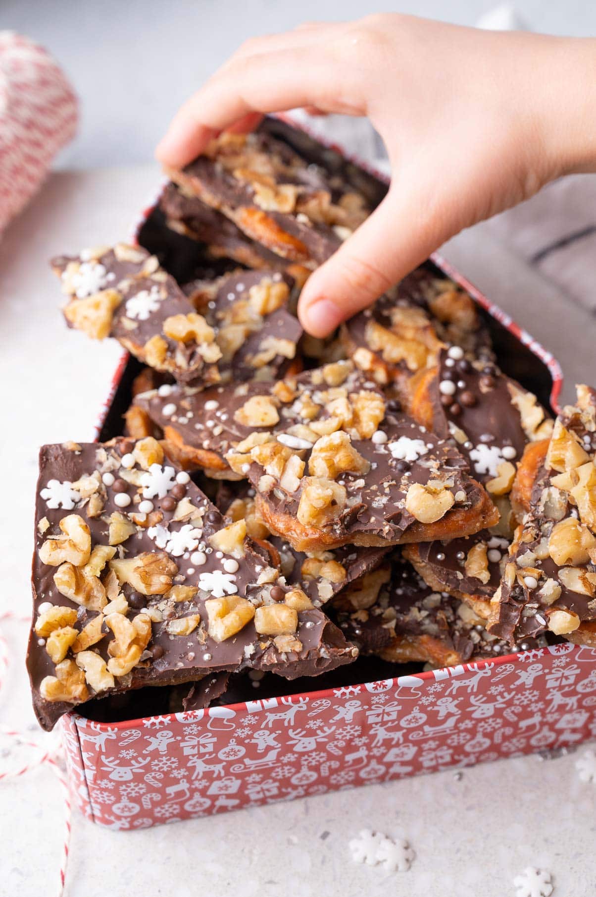 A child's hand is reaching for pretzel toffee in a cookie tin.