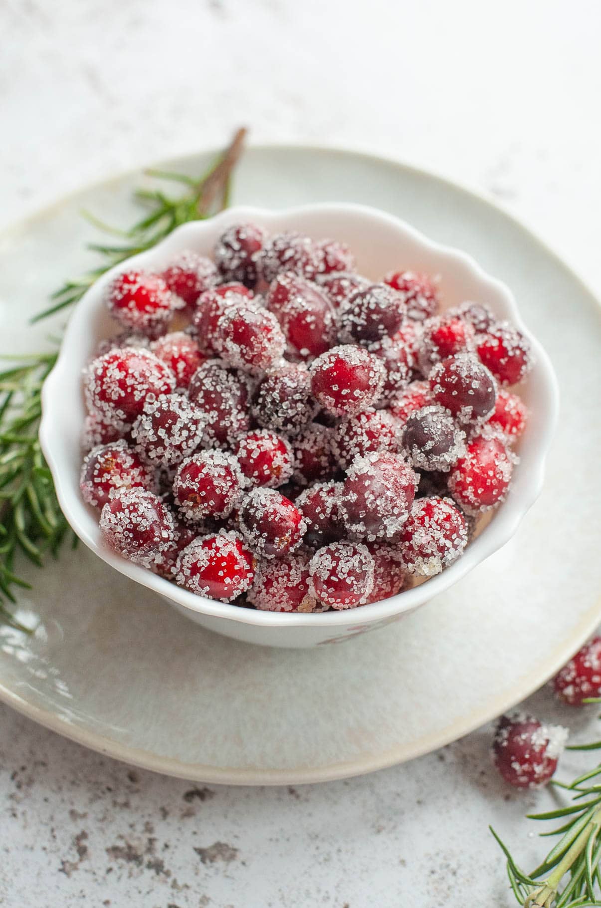Sugared cranberries in a white bowl, rosemary twigs in the background.