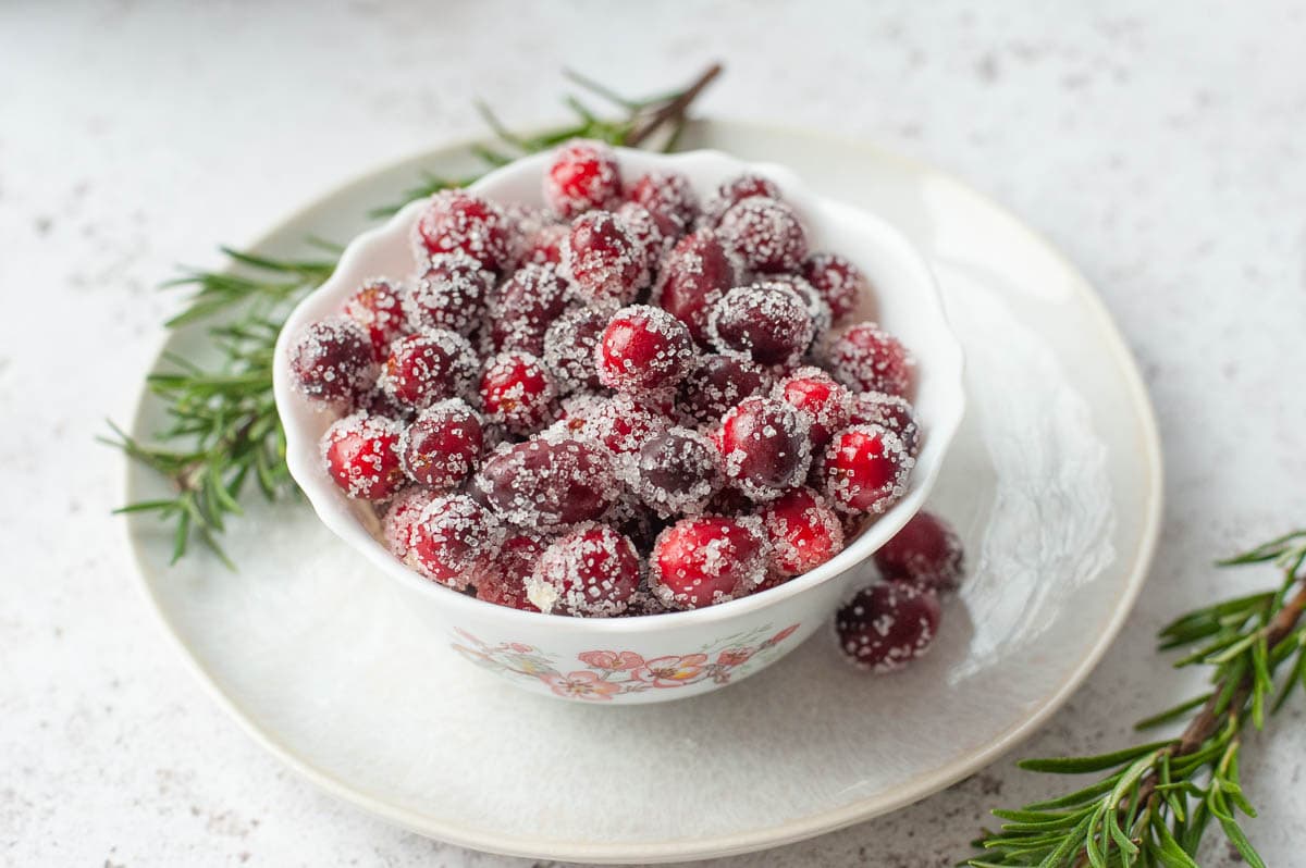 Sugared cranberries in a white bowl on a white plate. Rosemary twigs in the background.
