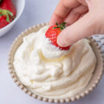 A strawberry is being dipped in whipped cream in a beige bowl.