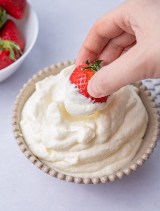 A strawberry is being dipped in whipped cream in a beige bowl.