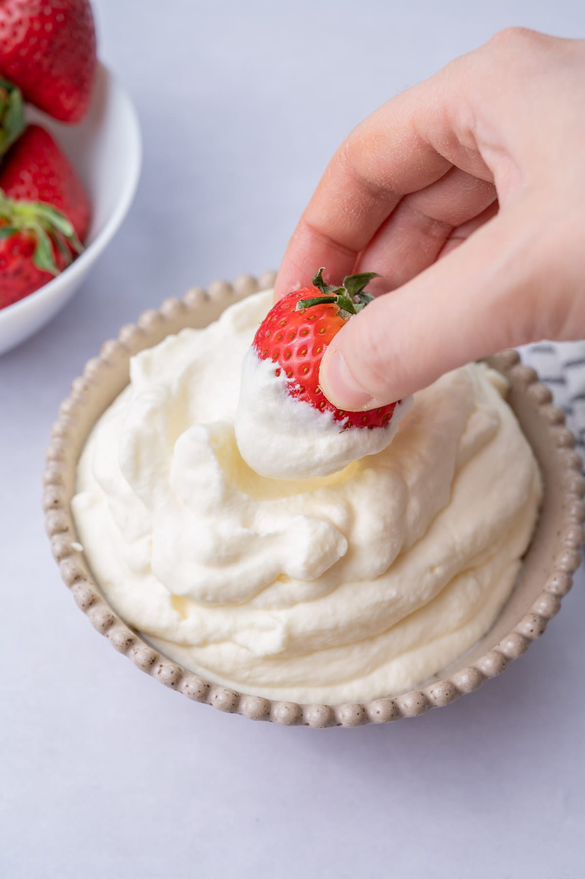 A strawberry is being dipped into whipped cream in a beige bowl.