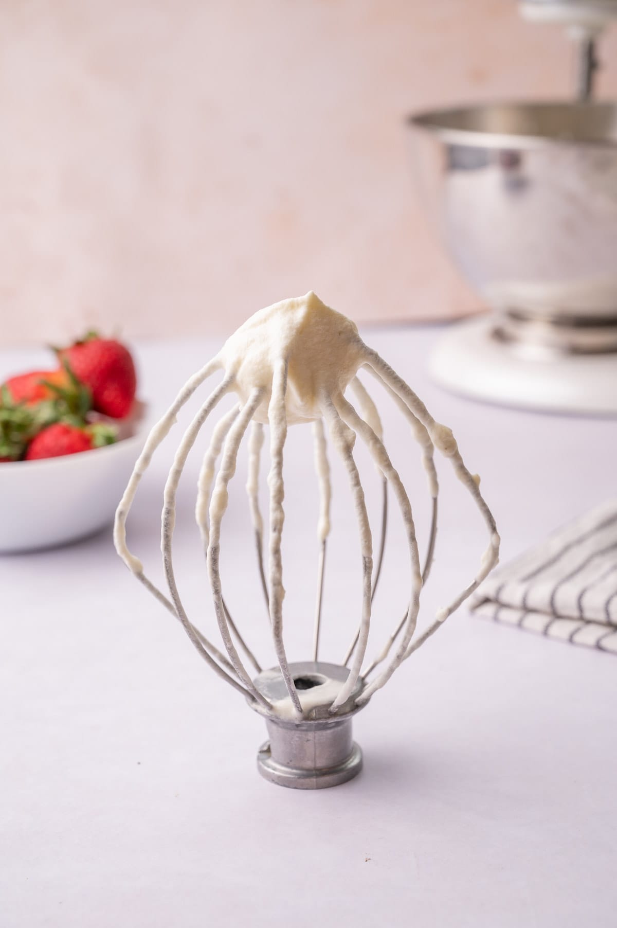 A mixer beater with whipped cream. Strawberries and a mixer in the background.