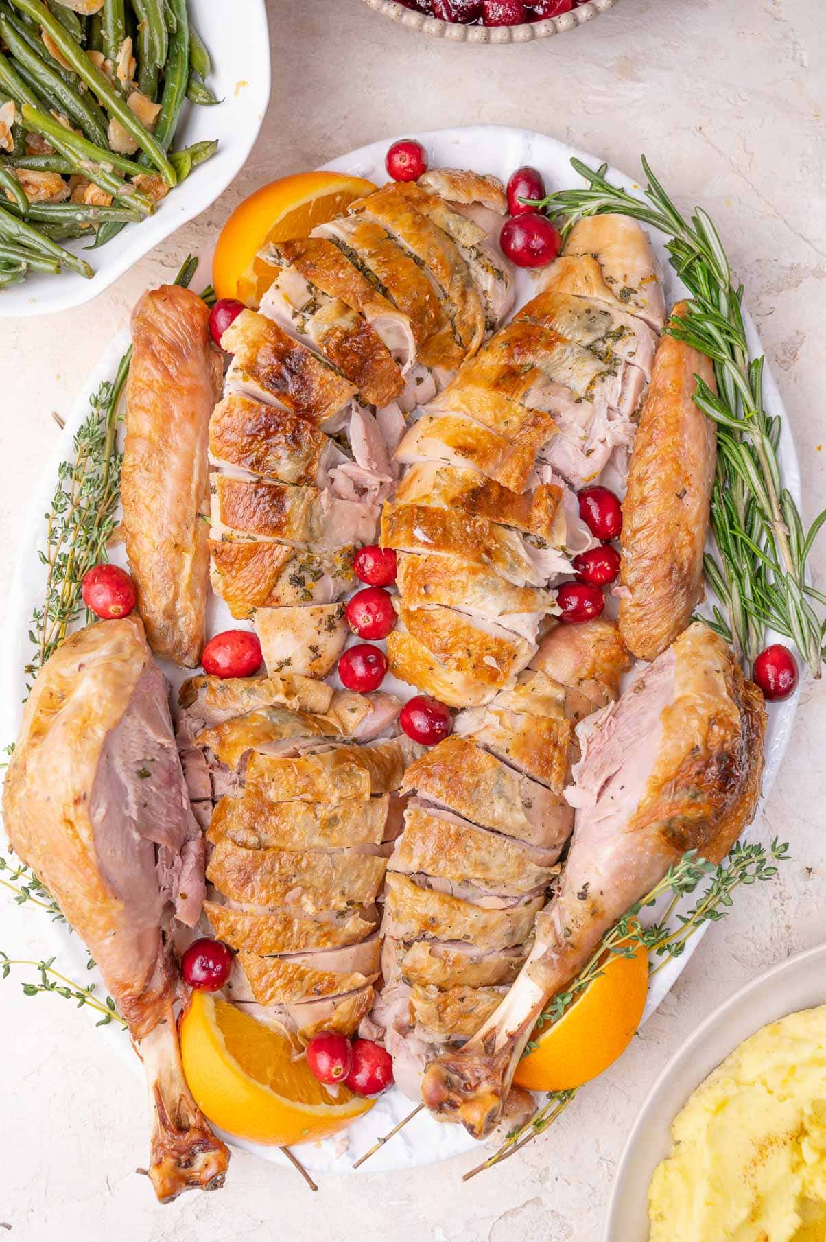 Carved turkey on a white plate surrounded with oranges, cranberries, and herbs.