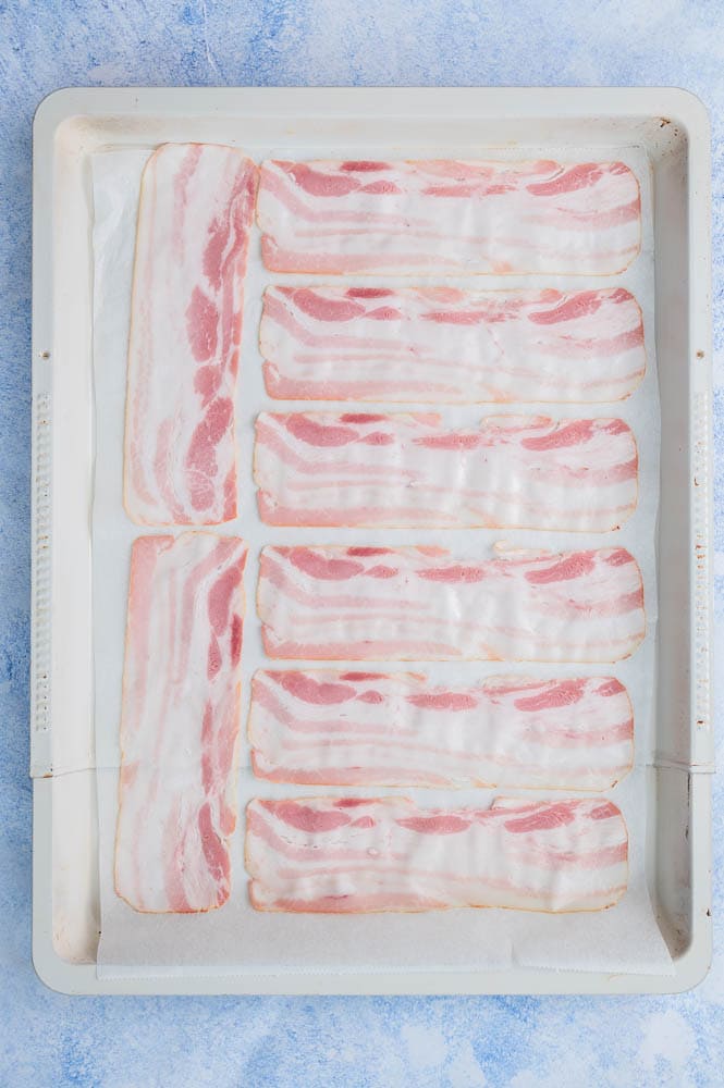 Raw bacon slices on a baking sheet lined with parchment paper.