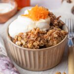 Carrot cake baked oatmeal in a brown bowl topped with cream cheese topping and grated carrots.