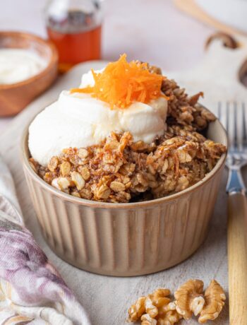 Carrot cake baked oatmeal in a brown bowl topped with cream cheese topping and grated carrots.