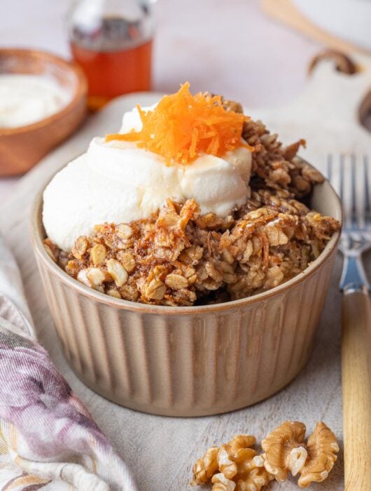 Carrot cake baked oatmeal in a brown bowl topped with cream cheese topping and grated carrots.