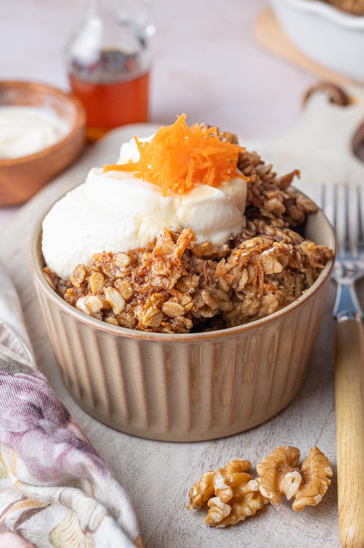 Carrot cake oatmeal in a brown bowl topped with cream cheese topping, grated carrots and walnuts.