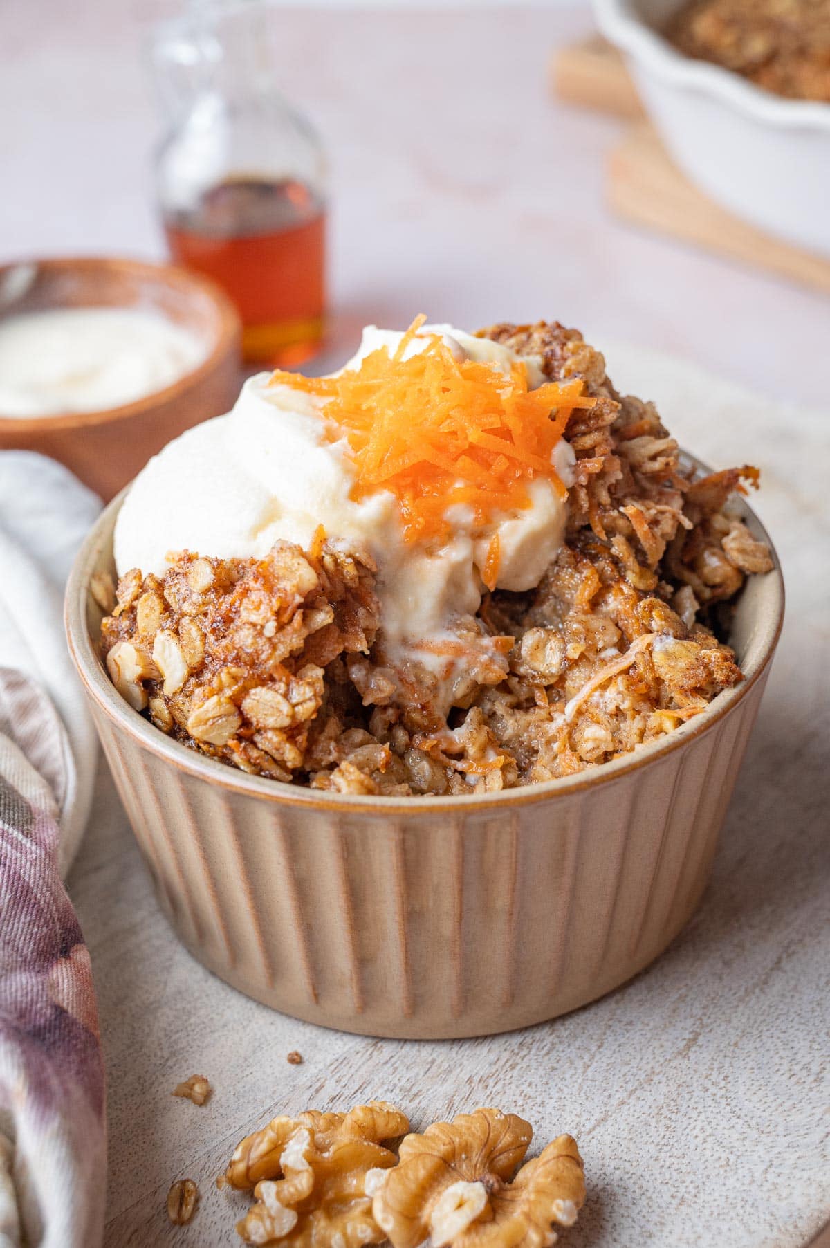 Carrot cake oatmeal in a brown bowl topped with cream cheese topping, grated carrots and walnuts.