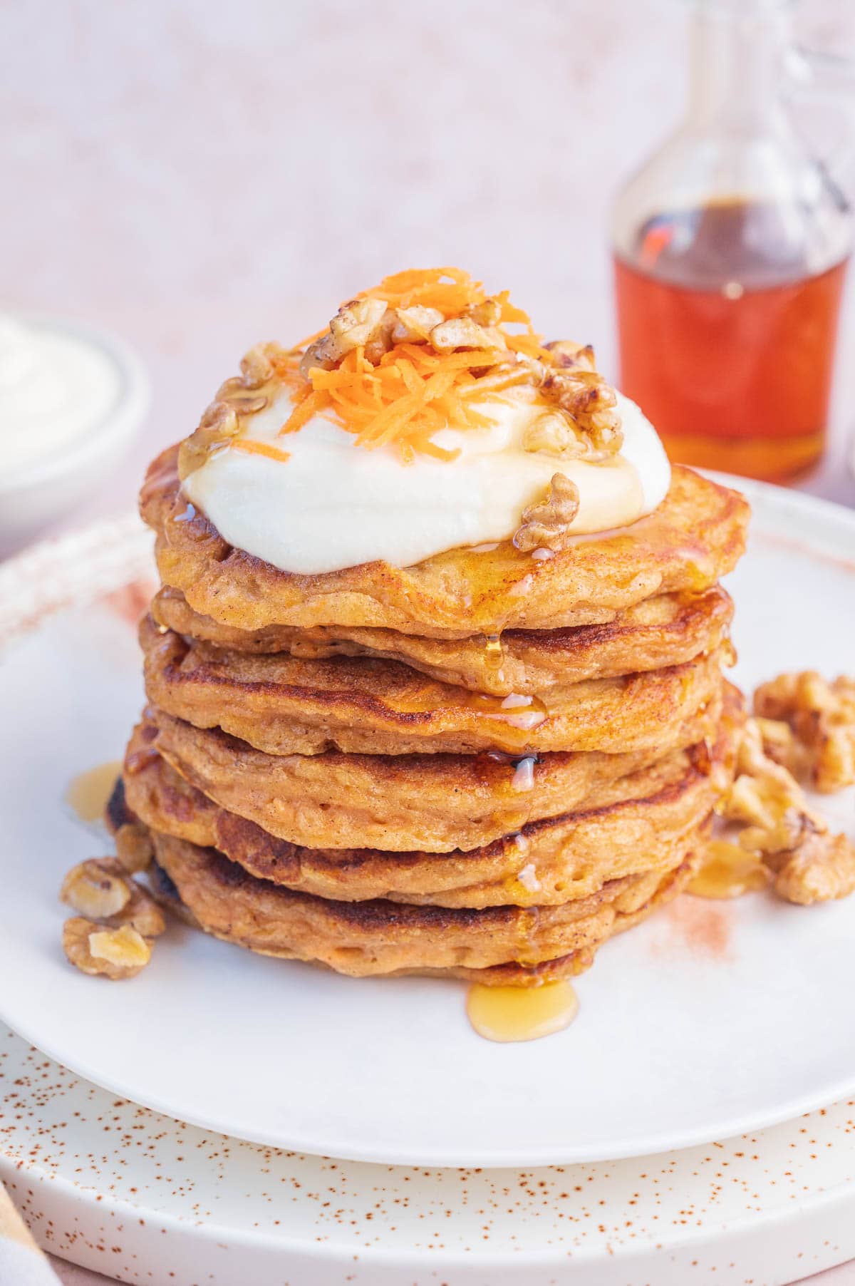 A stack of carrot cake pancakes on a white plate topped with cream cheese topping and walnuts.