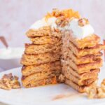 A stack of carrot cake pancakes on a white plate topped with cream cheese topping and walnuts.