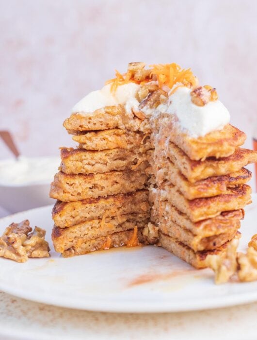 A stack of carrot cake pancakes on a white plate topped with cream cheese topping and walnuts.