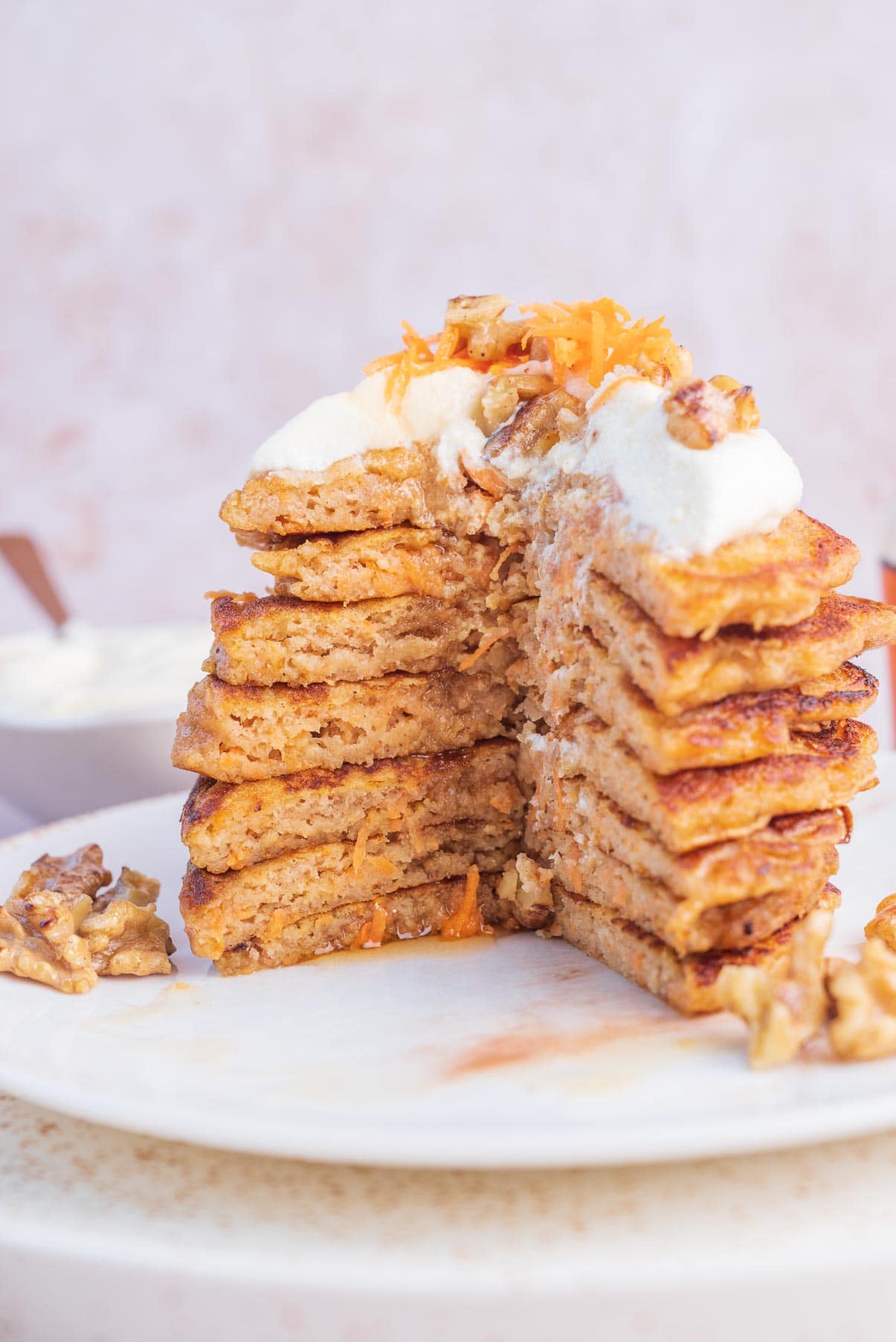 A stack of carrot cake pancakes with a part missing on a white plate topped with cream cheese topping and walnuts.