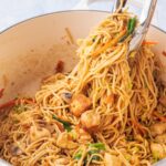 Chicken chow mein noodles in a white pot are being picked with kitchen tongs.