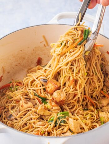 Chicken chow mein noodles in a white pot are being picked with kitchen tongs.