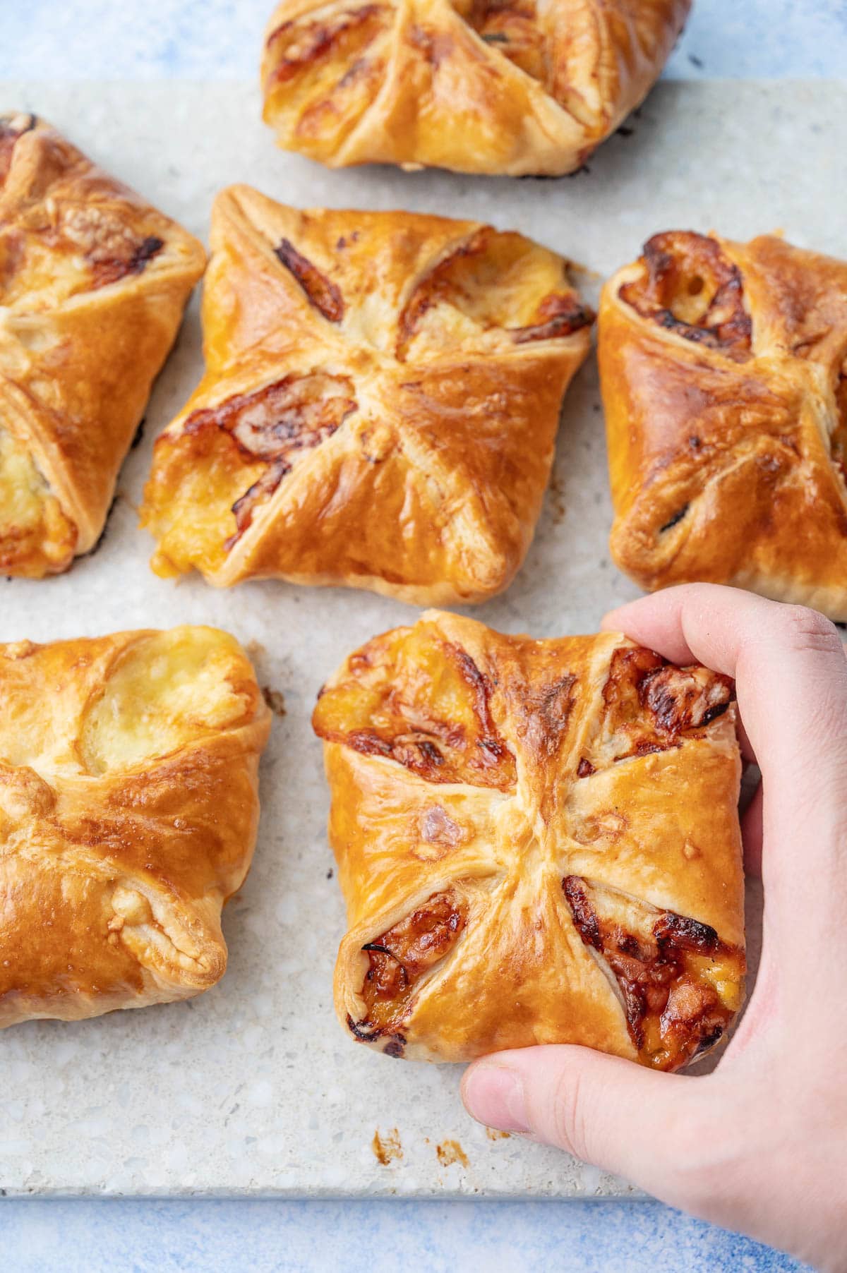 Ham and cheese puff pastry pastries on a grey stone board. One pastry is held in a hand.