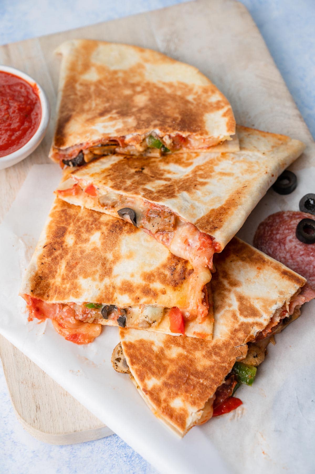 Pizza quesadillas on a white wooden board. A small bowl with pizza sauce in the background.