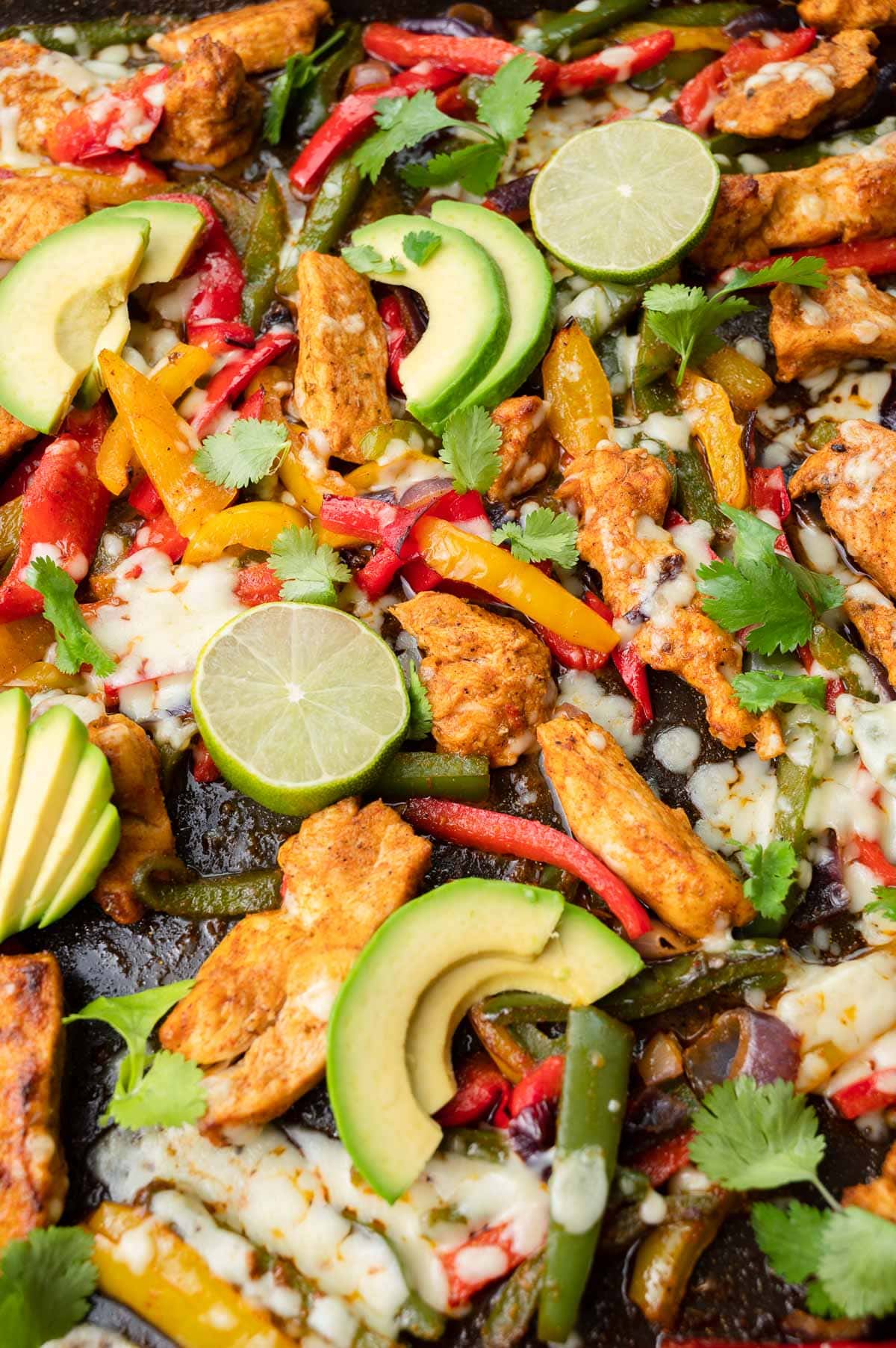 Sheet pan chicken fajitas on a black baking sheet topped with avocado slices, lime slices, and cilantro.