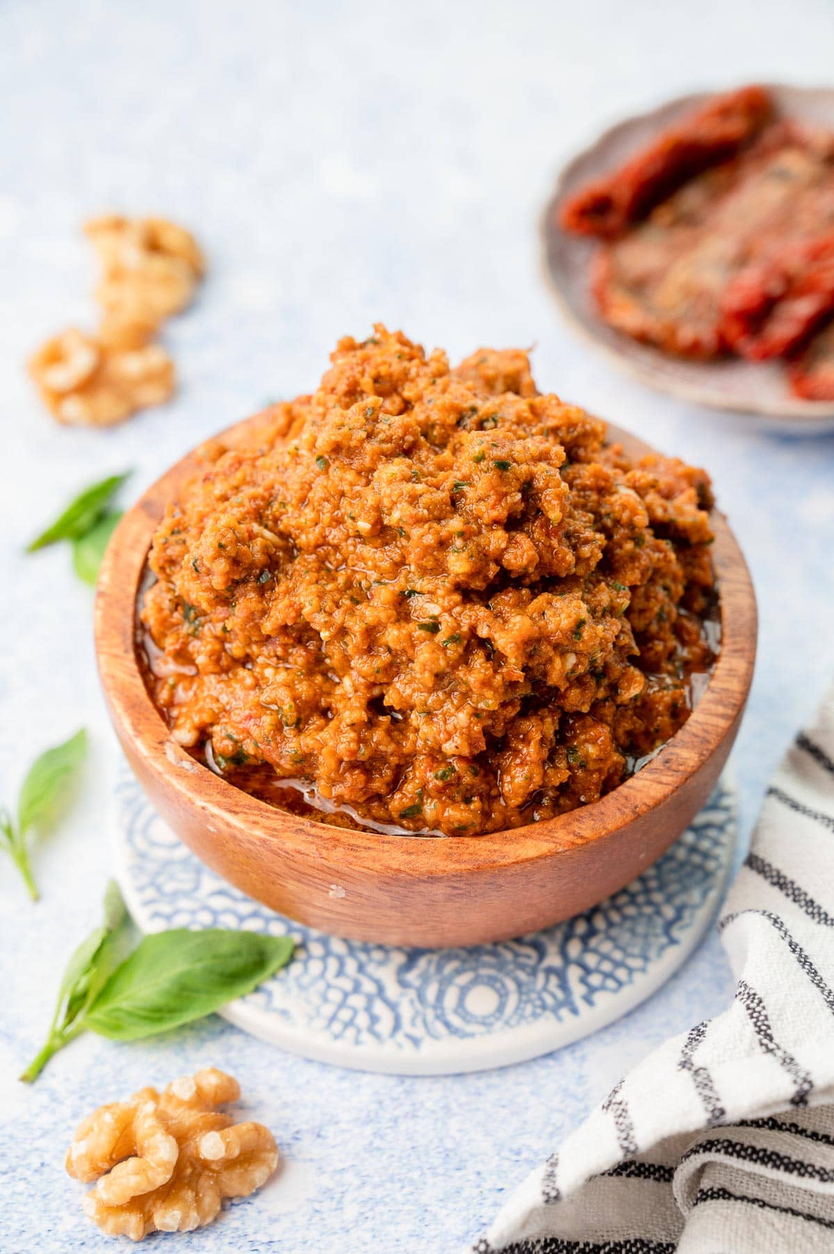 Sun-dried tomato pesto in a brown bowl. Basil leaves, sun-dried tomatoes and walnuts in the background.