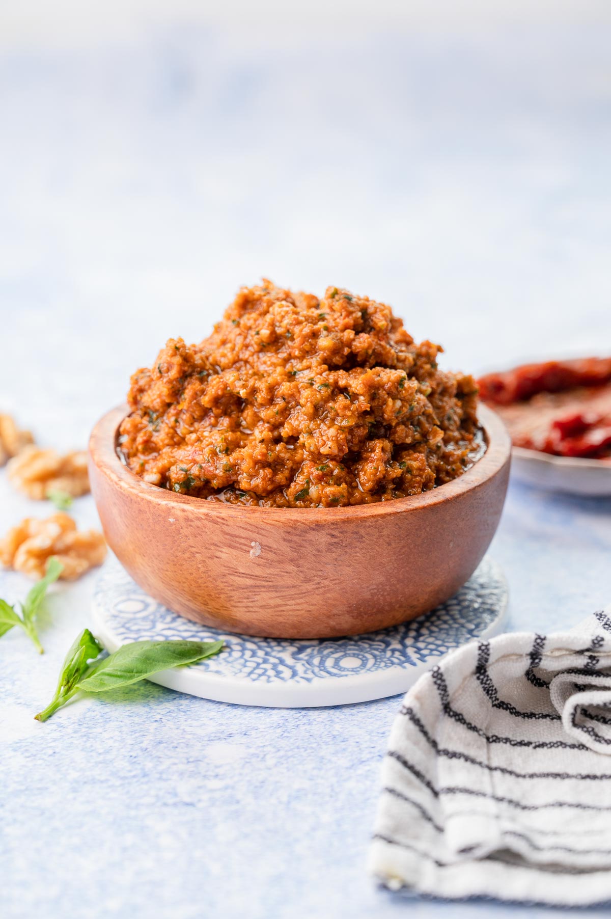 Red pesto in a brown bowl. Basil leaves, sun-dried tomatoes and walnuts in the background.