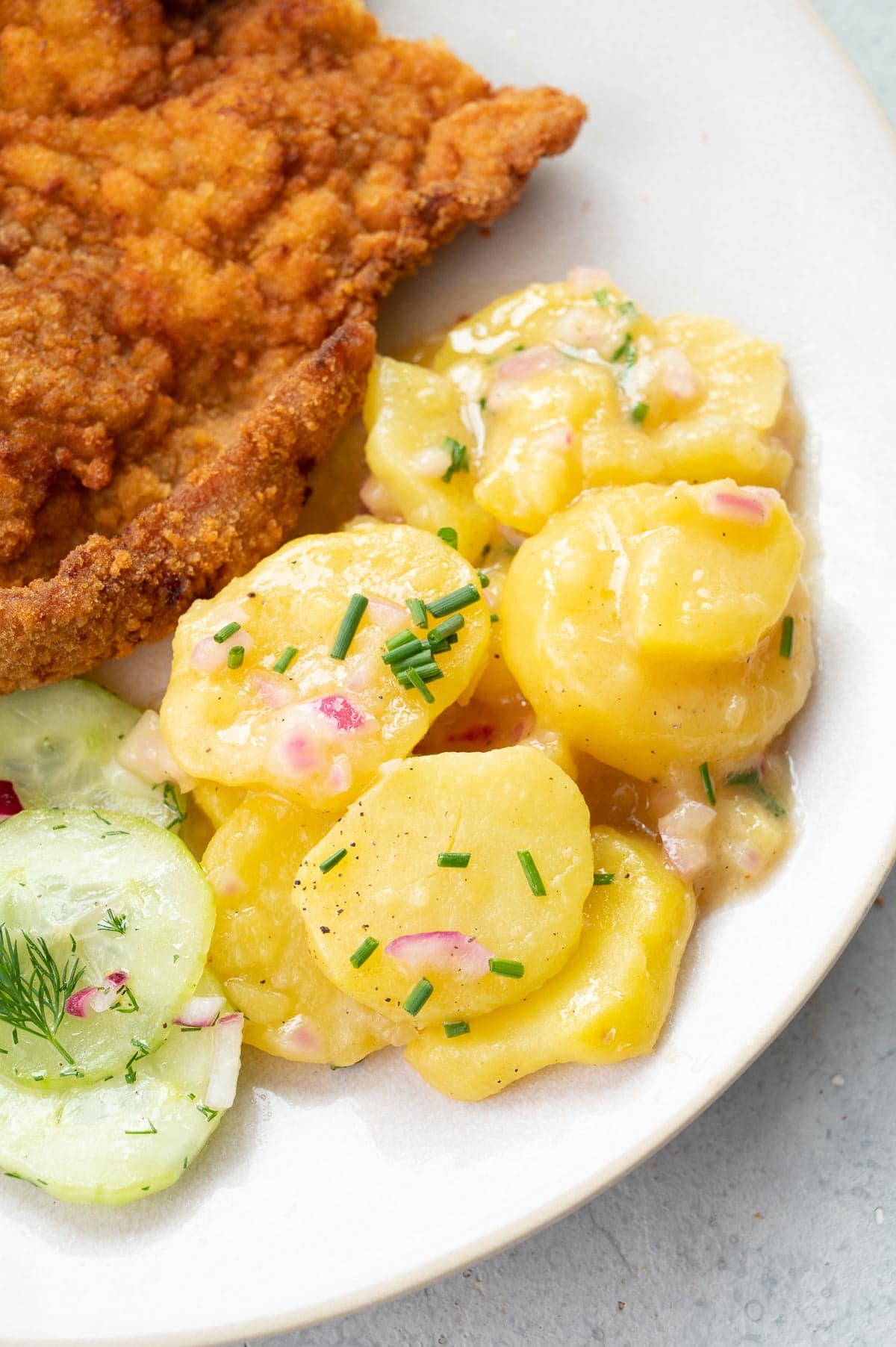 A close up photo of Austrian potato salad served with schnitzel and cucumber salad on a beige plate.