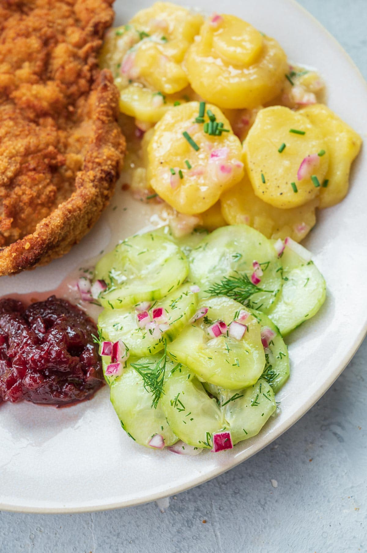 German cucumber salad on a beige plate with potato salad, Wiener Schnitzel and red currant preserves.