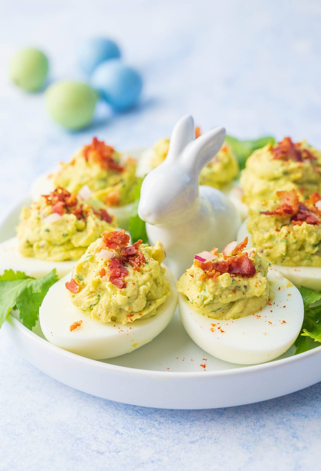 Avocado deviled eggs on a white plate topped with bacon and onions.