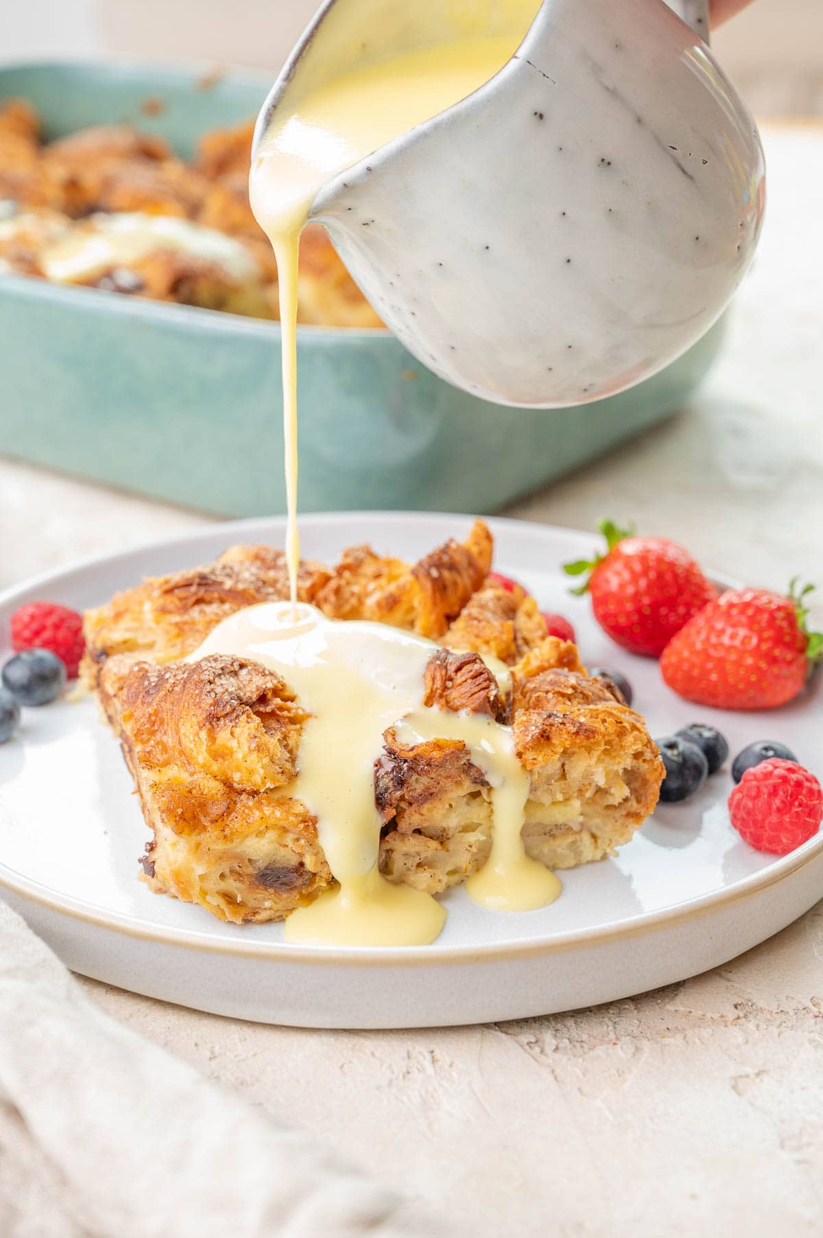 Vanilla sauce is being poured over croissant bread pudding on a white plate with berries.